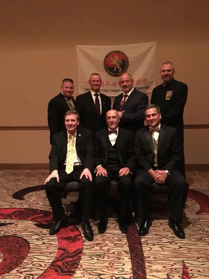 Men in dark suits standing in front of a flag at the ICHF 25th anniversary celebration.